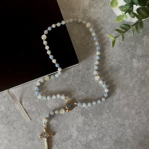 Op de afbeelding: Een rozenkrans met lichtblauwe en witte kralen, verbonden door goudkleurige metalen schakels. Het heeft een goudkleurige kruisvormige hanger en een cirkelvormig middelpunt met een religieus beeld. De rozenkrans ligt op een grijze ondergrond.