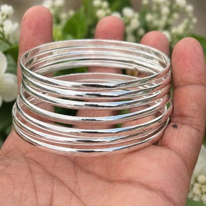 May include: A stack of shiny, silver bangles. The bracelets are round and smooth, reflecting light. The bangles are stacked on top of each other, creating a layered effect. The background is blurred, with hints of green and white.