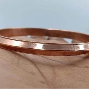 Puede incluir: Una pulsera de color cobre con un diseño plano y ancho. La pulsera tiene un acabado liso y pulido y un borde ligeramente redondeado. El metal refleja la luz. La pulsera es una joya sencilla y elegante. El diámetro es de aproximadamente 6,5 cm.