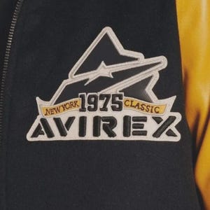 May include: Close-up of a black jacket with a white and black logo patch. The patch features the text "NEW YORK 1975 CLASSIC AVIREX" in a stylized font. The jacket has a yellow leather sleeve.