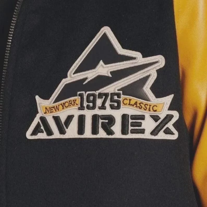 May include: Close-up of a black jacket with a white and black logo patch. The patch features the text "NEW YORK 1975 CLASSIC AVIREX" in a stylized font. The jacket has a yellow leather sleeve.