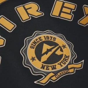 May include: Black fabric with yellow and white embroidered patches. The patches include the word "REX" and a circular emblem with "SINCE 1975 NEW YORK CLASSIC" text. The emblem features a lightning bolt design.