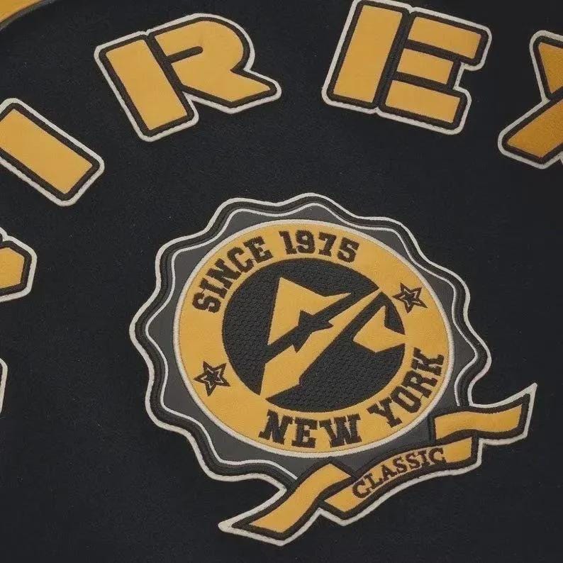 May include: Black fabric with yellow and white embroidered patches. The patches include the word "REX" and a circular emblem with "SINCE 1975 NEW YORK CLASSIC" text. The emblem features a lightning bolt design.