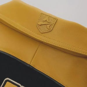 May include: Close-up of a mustard yellow leather item with a stitched emblem. The item has a black section at the bottom. The emblem is a shield shape with a detailed design.