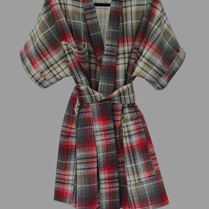 May include: A plaid dressing gown in red, grey, and white hangs on a black hanger. The robe has a wrap-around design with a matching belt. The fabric features a classic plaid pattern, ideal for relaxing.