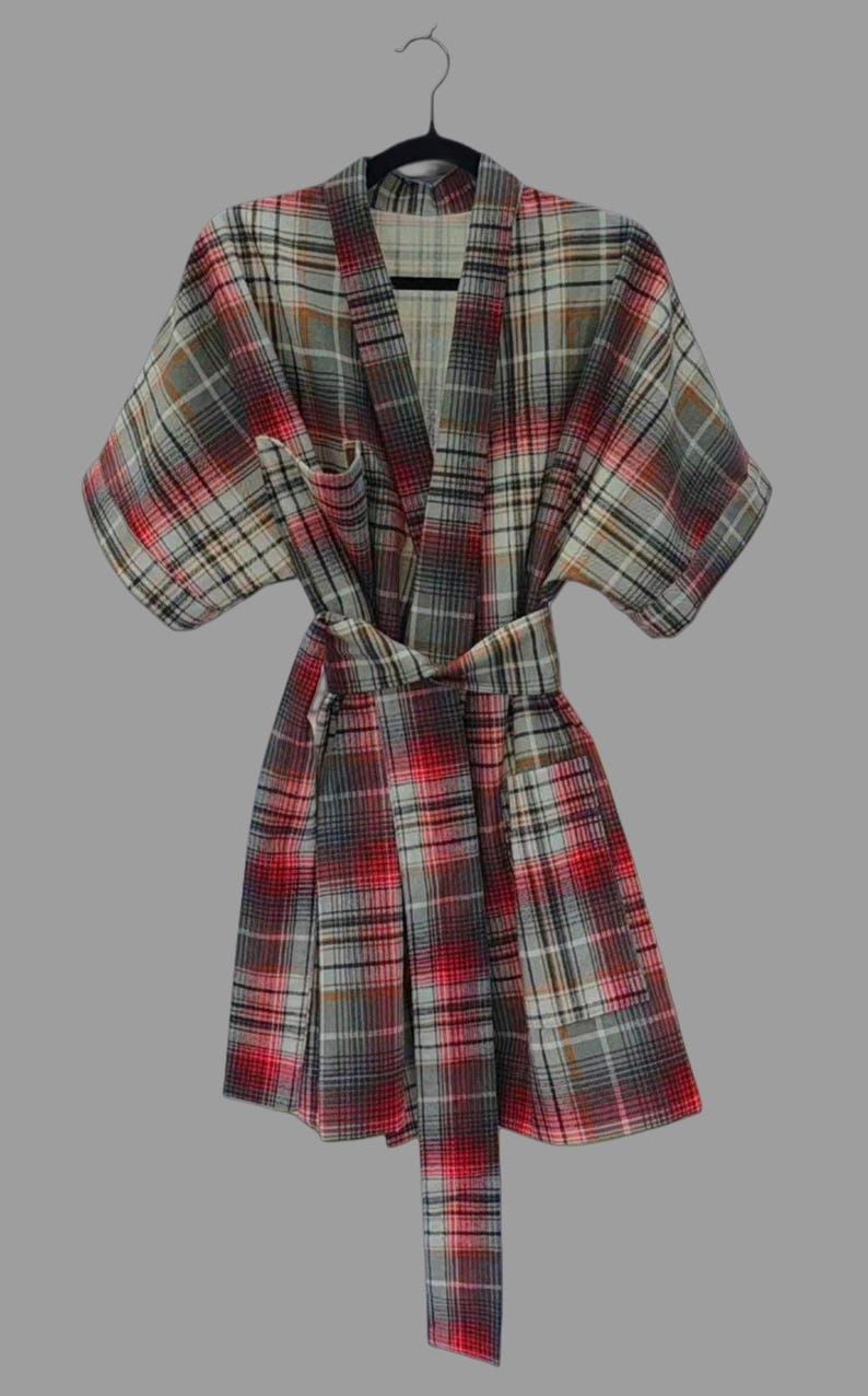 May include: A plaid dressing gown in red, grey, and white hangs on a black hanger. The robe has a wrap-around design with a matching belt. The fabric features a classic plaid pattern, ideal for relaxing.