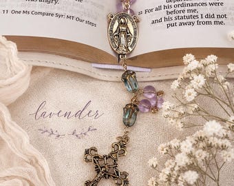 May include: A rosary featuring translucent light purple beads, a gold-toned metal centerpiece depicting a figure, and an ornate gold-toned crucifix. The rosary includes two blue-tinted floral-shaped beads and is placed on an open book page.