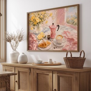Könnte beinhalten: Ein gerahmter Wandkunstdruck, der eine Frühstücksszene mit einer rosa Mokkakanne, einer Tasse Kaffee mit Latte Art, einem Croissant, Zitronenscheiben und einer Vase mit gelben Blumen auf einer rosa-weiß gemusterten Tischdecke zeigt.