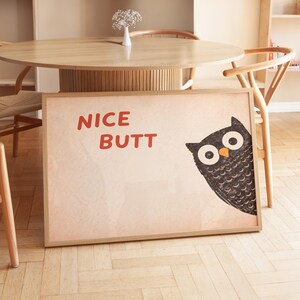 Könnte beinhalten: Ein gerahmter Druck mit dem Text "NICE BUTT" in Rot. Eine Cartoon-Eule mit großen Augen schaut von der rechten Seite hervor. Der Druck hat einen hellbraunen Hintergrund und einen Holzrahmen. Das Kunstwerk steht auf einem Holzboden.