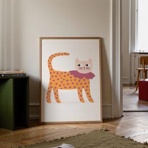 May include: A framed art print featuring a whimsical illustration of a cat. The cat has an orange body with purple polka dots, a pink face, and a ruffled collar. The artwork is set against a white background, framed in a light brown wooden frame.