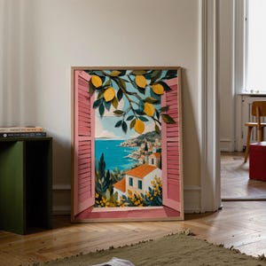 Könnte beinhalten: Ein rechteckiger gerahmter Kunstdruck mit einem Blick durch ein offenes Fenster mit rosa Fensterläden. Die Szene zeigt eine Küstenlandschaft mit weißen Gebäuden, orangefarbenen Dächern, blauem Wasser und einem Zitronenzweig mit gelben Früchten.