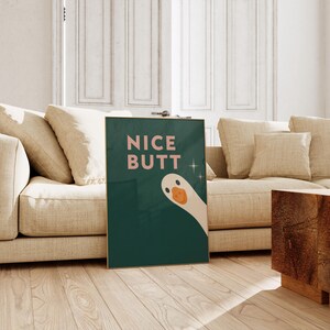 Könnte beinhalten: Ein rechteckiger Wandkunstdruck mit dunkelgrünem Hintergrund und dem Text NICE BUTT in hellrosa Großbuchstaben. Ein stilisierter cremefarbener Gänsekopf mit orangefarbenem Schnabel lugt aus der unteren rechten Ecke hervor, akzentuiert durch zwei kleine weiße vierzackige Sternsymbole.
