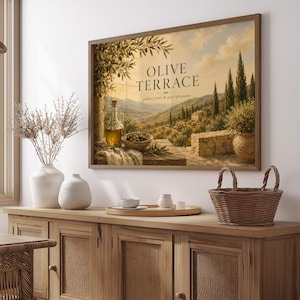 Könnte beinhalten: Ein gerahmter Landschaftsdruck mit dem Titel OLIVE TERRACE und dem Untertitel golden groves & quiet afternoons. Das Kunstwerk zeigt einen mediterranen Hang mit Olivenbäumen, einer Steinterrasse, einer Ölkaraffe aus Glas und einer Schale mit Oliven.