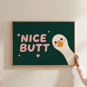 Könnte beinhalten: Ein rechteckiger Wandkunstdruck in einem Holzrahmen mit dunkelgrünem Hintergrund und dem rosa Text "NICE BUTT" in fetter Schrift. Das Design zeigt einen weißen Gänsekopf auf der rechten Seite, zwei kleine rosa Herzen und zwei weiße Sterne.