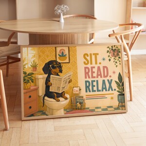 Könnte beinhalten: Ein gerahmter Kunstdruck mit einem schwarz-braunen Dackel mit gelber Brille, der auf einer Toilette sitzt und Zeitung liest. Der Text lautet SIT. READ. RELAX. mit Illustrationen von Topfpflanzen, einer Toilettenpapierrolle und einem Zeitschriftenständer.