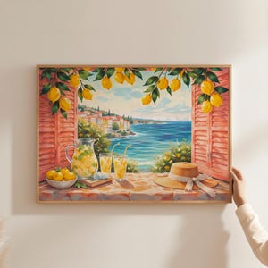 Könnte beinhalten: Ein gerahmter Kunstdruck, der den Blick aus einem offenen Fenster auf eine Küstenstadt und das blaue Meer zeigt. Die Szene umfasst einen Steinsims mit einer Karaffe Limonade, zwei Gläsern, einer Schale mit Zitronen und einem Strohhut.