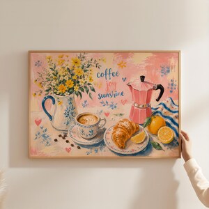 Könnte beinhalten: Ein gerahmter, rechteckiger Wandkunstdruck mit einer gemalten Szene aus einem weißen Krug mit gelben Blumen, einer Tasse Kaffee, einem Croissant, einer rosa Espressokanne und Zitrusfrüchten. Der Text coffee joy sunshine ist in Schreibschrift.