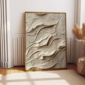 May include: A framed abstract art piece with a textured, layered design in shades of beige and cream. The artwork features a flowing, wave-like pattern, encased in a light brown wooden frame. The piece is leaning against a white wall.