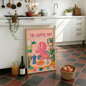 Könnte beinhalten: Ein gerahmter rechteckiger Druck mit einem rosa Triceratops, der an einem runden Tisch mit einer blauen Kaffeekanne, einem Croissant und einer Blumenvase sitzt. Der Text "TRI-COFFEE-TOPS" ist oben in Rosa gedruckt. Die Szene enthält grüne Pflanzen.