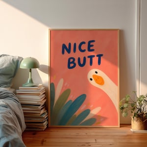 Könnte beinhalten: Ein gerahmter Kunstdruck mit dem Text "NICE BUTT" in blauen Buchstaben auf korallenfarbenem Hintergrund. Eine Cartoon-Gans schaut von der rechten Seite hervor. Grün-blaue Blätter akzentuieren die linke untere Ecke. Das Kunstwerk wird in einem Schlafzimmer gezeigt.