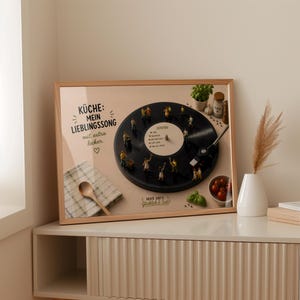 May include: A framed print featuring a black vinyl record with miniature figures arranged on it. The print includes text in German, a wooden spoon, and a small bowl of tomatoes. The frame is light brown.