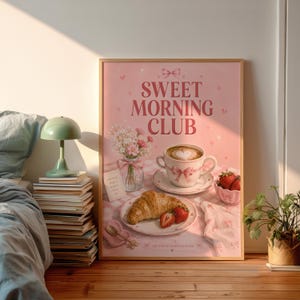 Könnte beinhalten: Ein rechteckiger Wandkunstdruck mit rosa Hintergrund und dem Text SWEET MORNING CLUB. Die Illustration zeigt eine Kaffeetasse mit herzförmigem Milchschaum, ein Croissant, frische Erdbeeren und eine Blumenvase auf einer karierten Tischdecke.