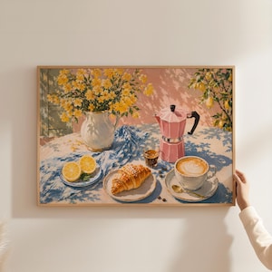 Könnte beinhalten: Ein gerahmtes Gemälde mit einem weißen Krug voller gelber Blumen, einer rosa Mokkakanne, einem Croissant auf einem Teller, einer Tasse Latte, einem kleinen Glas Espresso und Zitronenscheiben auf einem blau gemusterten Tuch vor einer rosa Wand.