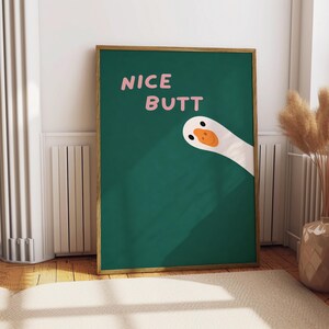 May include: A framed art print featuring a green background with the words "NICE BUTT" in pink. A cartoon goose peeks from the right side. The artwork is displayed on a wooden floor.