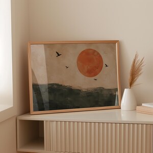Puede incluir: Una impresión de arte mural enmarcada rectangular que presenta un paisaje con un sol grande y naranja sólido sobre un cielo beige apagado. Cuatro siluetas de pájaros negros vuelan sobre una línea de horizonte verde oscuro texturizada.