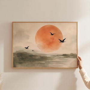 May include: A rectangular wall art print in a light wood frame featuring a watercolour landscape. The scene depicts a large, muted orange sun centred in a hazy sky above dark, abstract green-grey hills with four black birds flying across.