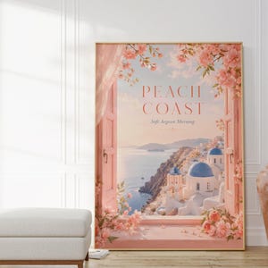 Könnte beinhalten: Ein gerahmter Kunstdruck mit dem Titel "Peach Coast" zeigt eine Küstenlandschaft mit weißen Gebäuden, blauen Kuppeln und rosa Blumen. Das Kunstwerk befindet sich in einem goldenen Rahmen und ist vor einer weißen Wand platziert. Der Druck hat eine weiche, verträumte Ästhetik.