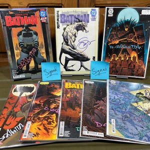 May include: A collection of Batman comic books, including several signed editions. The covers feature various illustrations of Batman and related characters, with a range of color palettes and artistic styles. Some comics have the word "Signed" on a blue sticky note.