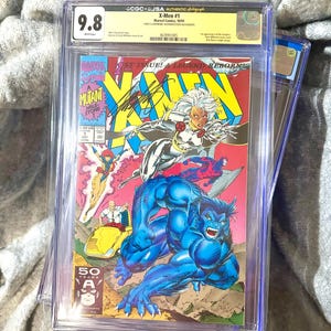 May include: A graded X-Men #1 comic book, featuring the characters Storm and Beast, with a grade of 9.8. The cover art is vibrant with a yellow, red, and blue color scheme. The comic is encased in a protective plastic sleeve.