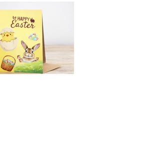 Op de afbeelding: Een gele Paas wenskaart met een cartoon illustratie van een kuiken dat uit een ei komt, een bruin konijn en een mand met eieren. De woorden "Be Happy Easter" staan bovenaan de kaart.