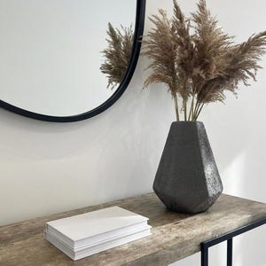 Puede incluir: Una escena interior moderna con un jarrón geométrico negro lleno de hierba de la pampa seca. Una pila de libros blancos descansa sobre una consola de madera. Un espejo redondo con marco negro cuelga de la pared blanca.