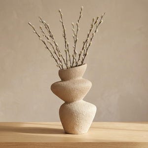May include: A beige, textured ceramic vase with a unique stacked design, holding several branches of pussy willow. The vase is on a light wood surface, with a neutral background.