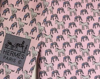 May include: A light pink necktie featuring a repeating pattern of small white horses and blue-topped gymnastic pommel horses. A dark gray rectangular label is stitched onto the fabric, displaying the text HERMÈS PARIS with a horse-drawn carriage icon.