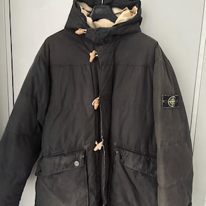Può includere: Un parka con cappuccio color grigio antracite scuro caratterizzato da una chiusura frontale con alamari in legno chiaro, due ampie tasche con patta in vita e un logo circolare a forma di bussola applicato sulla manica sinistra. La fodera è beige.
