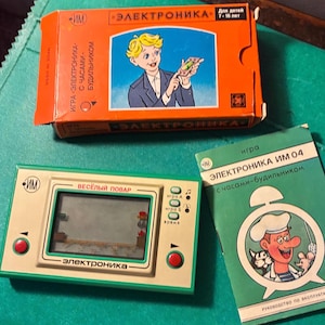 Pode incluir: Um jogo eletrônico portátil vintage Elektronika com um console bege e verde com botões vermelhos, uma caixa de papelão laranja com a ilustração de um menino e um manual de instruções verde com um chef, tudo com texto em cirílico.