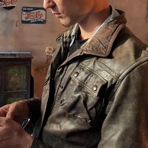 May include: A dark brown leather jacket featuring a pointed collar, two chest pockets with snap button closures, and a visible plaid lining at the neckline. The jacket has a textured, worn-in appearance with visible stitching and metal hardware.