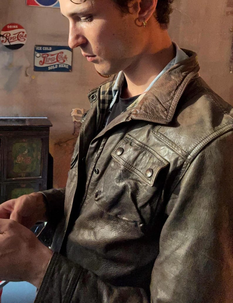 May include: A dark brown leather jacket featuring a pointed collar, two chest pockets with snap button closures, and a visible plaid lining at the neckline. The jacket has a textured, worn-in appearance with visible stitching and metal hardware.