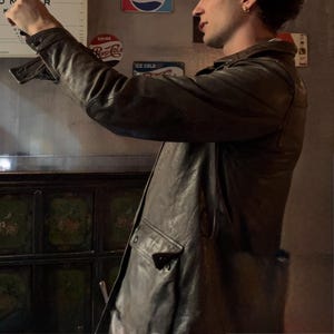 May include: A dark brown leather jacket featuring a structured collar, long sleeves with buttoned cuffs, and a prominent front pocket with a flap closure. The jacket displays a smooth, slightly reflective texture and is captured from a side profile.