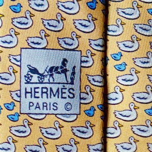 Könnte beinhalten: Eine goldfarbene Krawatte mit einem sich wiederholenden Muster aus weißen Enten mit blauen Akzenten und gelben Schnäbeln. Ein quadratisches Etikett ist mittig auf dem Stoff platziert und zeigt eine schwarze Kutsche über dem Text HERMÈS PARIS.