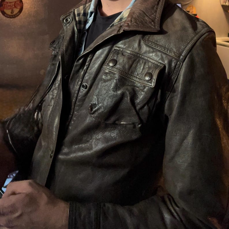 May include: A dark brown leather jacket featuring a folded collar, a front zipper closure with snap buttons, and two chest pockets with snap-button flaps. The interior lining displays a tan, black, and red plaid pattern visible at the neckline.