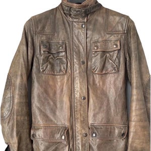 May include: A distressed brown leather jacket featuring a high collar with a buckle strap, a front button placket, two chest pockets with buttoned flaps, and two lower pockets with buttoned flaps. The jacket includes reinforced elbow patches.