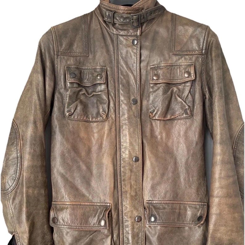 May include: A distressed brown leather jacket featuring a high collar with a buckle strap, a front button placket, two chest pockets with buttoned flaps, and two lower pockets with buttoned flaps. The jacket includes reinforced elbow patches.