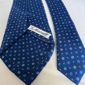 Könnte beinhalten: Eine dunkelblaue Krawatte mit einem sich wiederholenden Muster aus kleinen hellblauen Blumen und winzigen violetten ovalen Formen. Ein weißes Stoffetikett ist auf der Rückseite der Krawatte aufgenäht und zeigt den schwarzen Text E. Marinella NAPOLI.