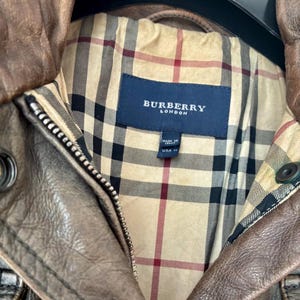 May include: A brown leather jacket featuring a beige, black, and red plaid interior lining. A dark blue rectangular label with the white text BURBERRY LONDON is stitched inside, along with a smaller tag indicating Made in Italy and USA size 14.