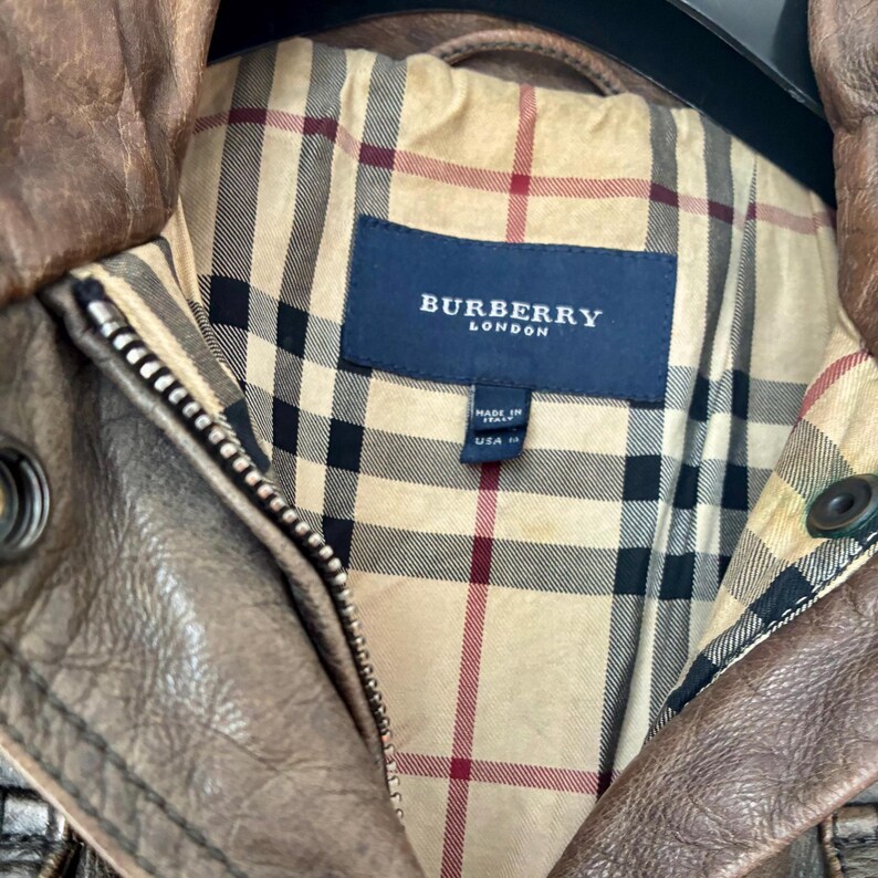 May include: A brown leather jacket featuring a beige, black, and red plaid interior lining. A dark blue rectangular label with the white text BURBERRY LONDON is stitched inside, along with a smaller tag indicating Made in Italy and USA size 14.