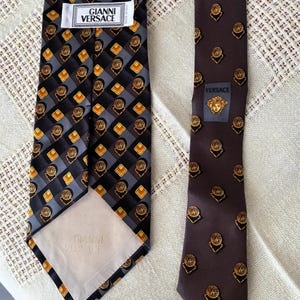 May include: Two silk neckties. One is black with a geometric pattern of gray, gold, and black squares and circles. The other is brown with a repeating gold medallion design. Both ties have the "Gianni Versace" label.